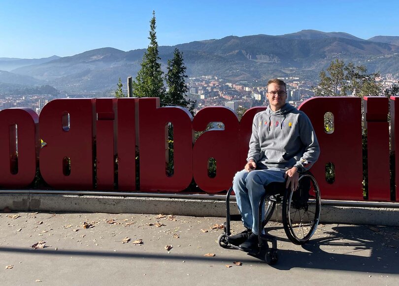Bild von Lukas Gloßner in Jeans und Hoody im Rollstuhl vor dem Schriftzug Bilbao im Vordergrund vor dem Panorama von Bilbao im Hintergrund
