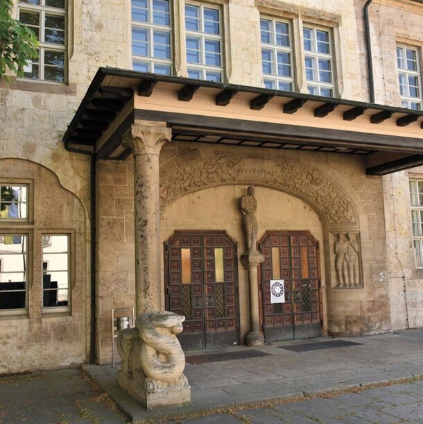 Eingangsportal zum historischen Hauptgebäude der Uni Jena mit Säulen und Fresken.