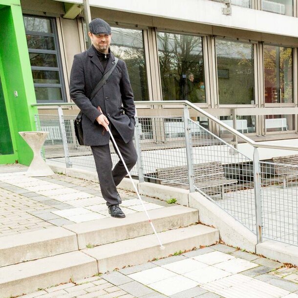 2.	Ein sehbehinderter Mann mit einem Blindenstock tritt aus einem Gebäude der TU Dortmund; er nutzt zur Orientierung das taktile Leitsystem auf dem Boden, das an vielen Stellen der TU vorhanden ist.