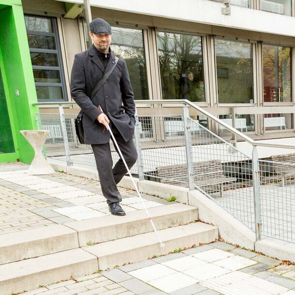 A visually impaired man with a cane steps out of a building of the TU Dortmund; he uses the tactile guidance system on the floor, which is available in many places of the TU, for orientation.