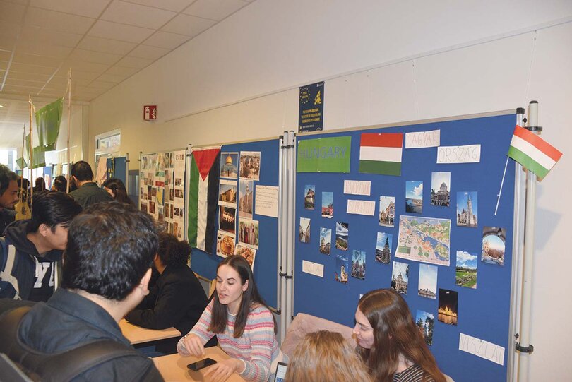Blue display walls with information on various countries can be seen in one of the university buildings. Students sit in front of them, appearing to answer questions from other students who are taking advantage of the "International Village" programme.